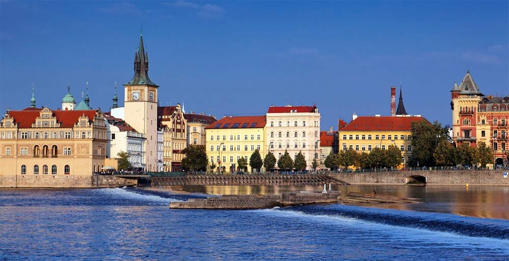 Panoramisk udsigt over Prags skyline ved floden, med historiske bygninger og en klar blå himmel, illustrerer byens charme og kulturelle arv.